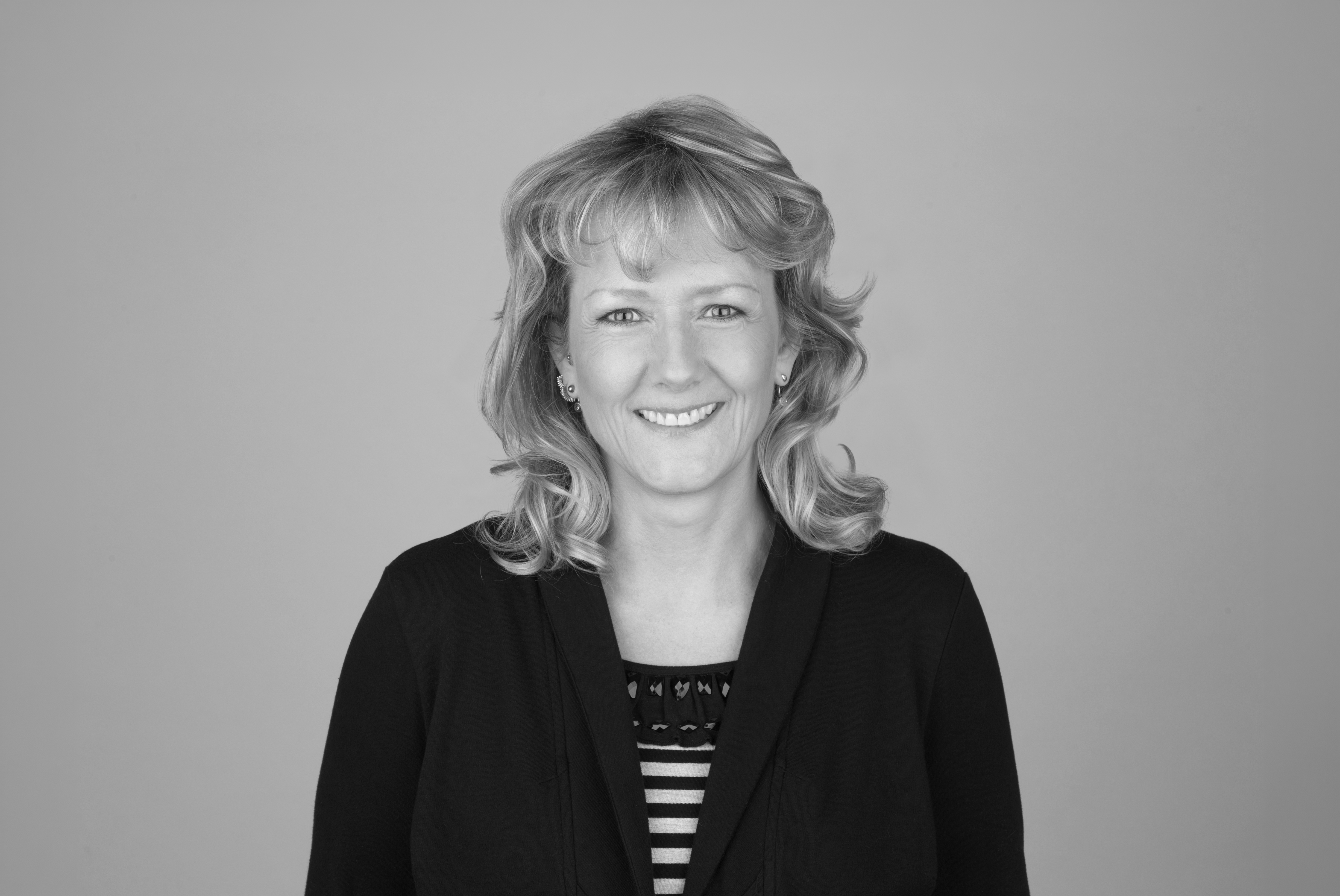 Eine lächelnde Frau mit blonden, leicht gewellten Haaren trägt einen schwarzen Pullover über einem gestreiften Oberteil und steht vor einem grauen Hintergrund.