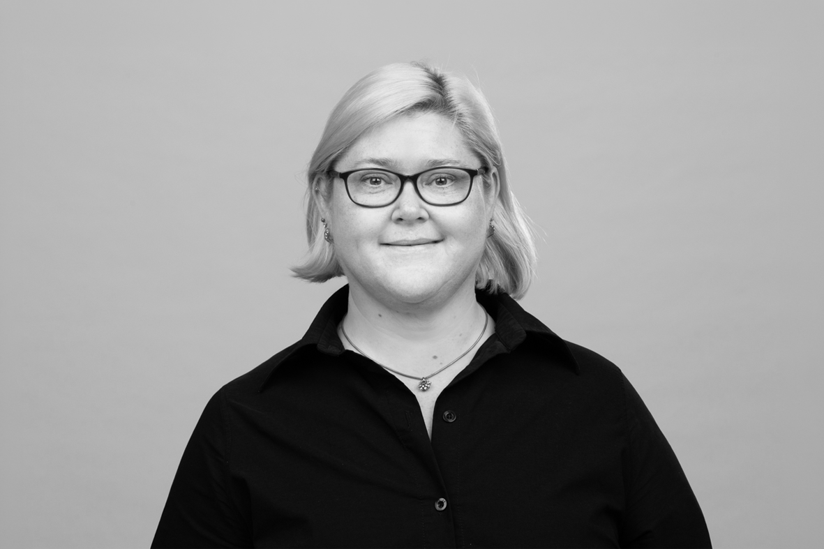 Eine lächelnde Person mit blonden, schulterlangen Haaren und einer schwarzen Bluse steht vor einem grauen Hintergrund. Sie trägt eine Brille und hat eine entspannte Ausstrahlung.